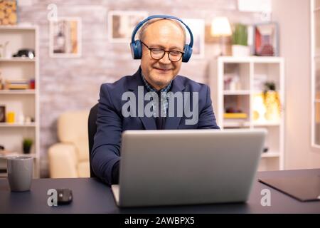 Ältere Mann in seinem 60s mit Kopfhörern auf dem Kopf Musik hören und arbeiten auf einem modernen Laptop Stockfoto