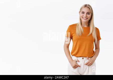 Selbstbewusste, blonde, stilvolle skandinavische Frau in orangefarbenem T-Shirt, hält die Hände an der Hose und lächelt selbstsicher, fühlt sich ermutigt, den Starttag zu beginnen Stockfoto