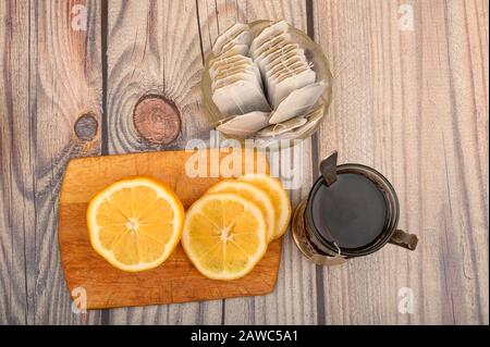 Tee aus facettiertem Glas in einem Vintage-Cup-Halter, Teebeutel in einer Glasvase und geschnittene Zitrone auf einem Brett auf Holzhintergrund. Nahaufnahme Stockfoto