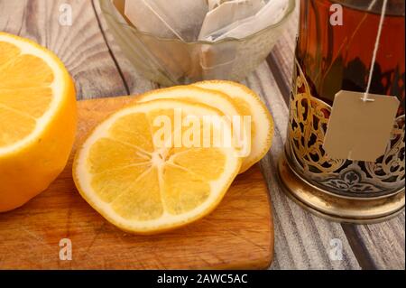 Tee aus facettiertem Glas in einem Vintage-Cup-Halter, Teebeutel in einer Glasvase und geschnittene Zitrone auf einem Brett auf Holzhintergrund. Nahaufnahme Stockfoto