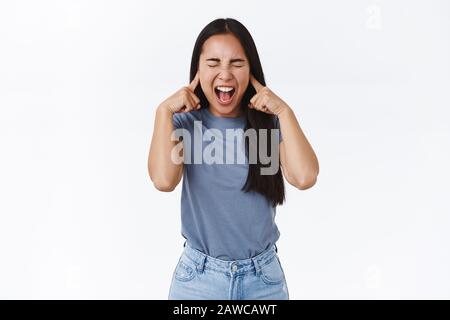 Verärgert und gefüttert asiatischen Teenager-Mädchen mit langen dunklen Haaren, schreiend verlorenem Temperament, kann nicht die Emotionen kontrollieren, Augen schließen und kreischen, Ohren schließen mit Stockfoto