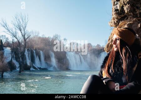 Attraktive Brunette, die auf einer Wand mit Blick in die Ferne posiert. Kravice-Wasserfall in der Ferne, bosnien und herzegowina. Kalter Wintertag Stockfoto