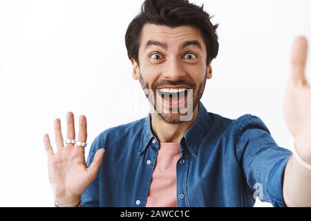 Nahestehender, freundlicher junger Mann mit Borste, dunkler modischer Frisur, ziehen Sie die Hand nach vorne, nehmen sie selfie auf das Smartphone, sprechen Sie per Videoanruf und Stockfoto