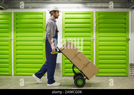 Seitenansicht des jungen Mannes in Overalls mit Wagen mit Kisten entlang des Korridors des Lagers Stockfoto