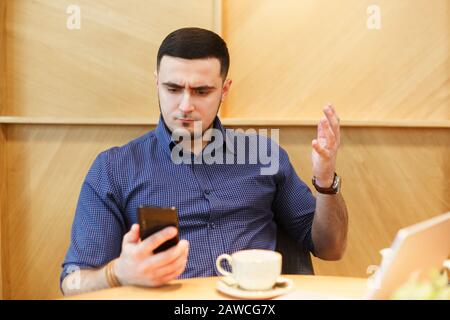 Der wütende Mann sieht beim Sitzen in einem Café auf den Telefonbildschirm und verstickt mit der Hand. Er ist mit etwas unzufrieden Stockfoto