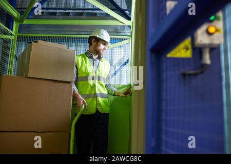 Junger Arbeiter in Arbeitskleidung und in Arbeitshelmen, der im Aufzug steht, mit Last auf dem Wagen Stockfoto