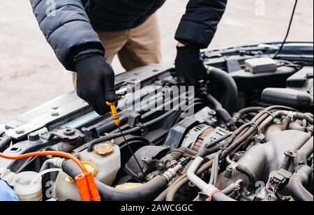 Mann mit schwarzen Handschuhen, der den Ölstand im Auto im Winter im Freien überprüft. Kfz-Mechaniker, der im Kfz-Reparaturdienst arbeitet. Stifthände zur Befestigung eines Autos Stockfoto