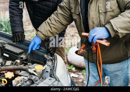 2 Mechaniker laden Autobatterie mit Strom über Überbrückungskabel im Freien auf. Rote und schwarze Überbrückungskabel in den männlichen Händen des Kfz-Mechanikers Stockfoto