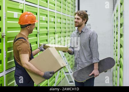 Junger Arbeiter im Arbeitshelm, der jungen Mann ein Paket gibt, während sie im Lager stehen Stockfoto