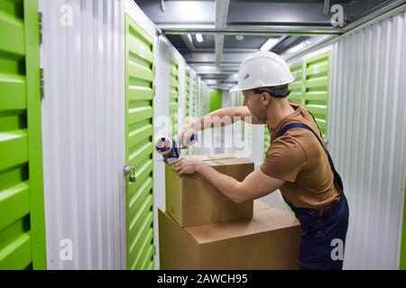 Junger Mann in Arbeitshelm-Packkästen mit Klebeband und in die Lagerkästen im Lager Stockfoto