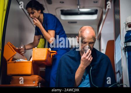 Der Mensch atmet durch die Sauerstoffmaske, die junge Krankenschwester in medizinischer Uniform öffnet etwas, das aus der medizinischen Tasche im Rettungswagen genommen wurde Stockfoto