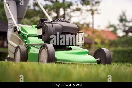 Nahaufnahme der weiblichen Beine des Gärtners mit grünem Rasenmäher auf dem Hinterhof. Selektiver Fokus der Frau, die im Sommer arbeitet und Gras im Hinterhof schneidet. Konzept Stockfoto