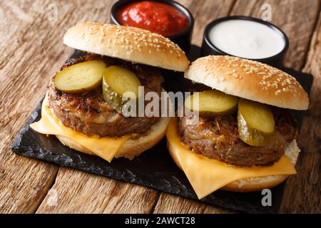 Hausgemachte gebratene Zwiebelburger mit Cheddar-Käse und Pickeln liegen auf einem Schieferbrett auf einem Tisch. Horizontal Stockfoto