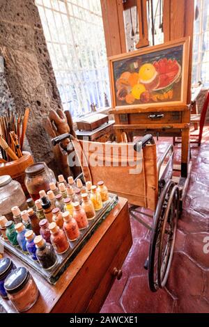 Farbflaschen stehen hinter dem Rollstuhl von Frida Kahlo im Casa Azul Museum in Coyoacan, CDMX, Mexiko. Stockfoto