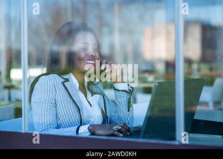 Schöne afroamerikanische Frau, die einen Laptop benutzt und auf ihrem Handy hinter einem Fenster spricht Stockfoto