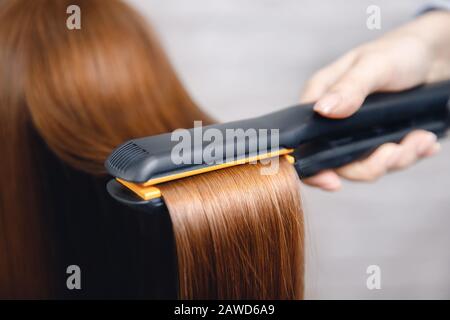 Mädchen glättet lange glänzend rote Haare mit Eisen im Schönheitssalon Stockfoto