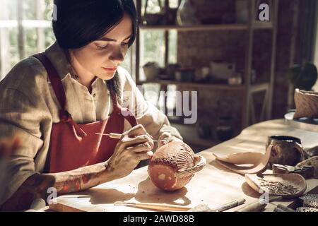 Craftsperson Konzept. Junge Frau, die im Kreativstudio Töpferwaren anstellt, um Details über das Sitzen der Tasse mit dem Modellierungstool in der dunklen, konzentrierten Umgebung zu erstellen Stockfoto