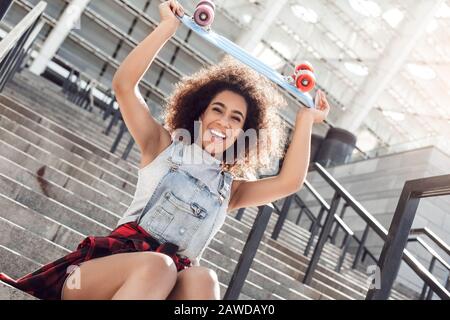 Junge Frau in der Straße der Stadt sitzt auf einer Treppe mit Penny Board sieht Kamera verwirrt aus Stockfoto