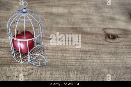 Rotes Herz in einem Vogelkäfig auf einem Holztisch. Selektive Fokussierung auf die offene Tür. Kopierbereich. Stockfoto