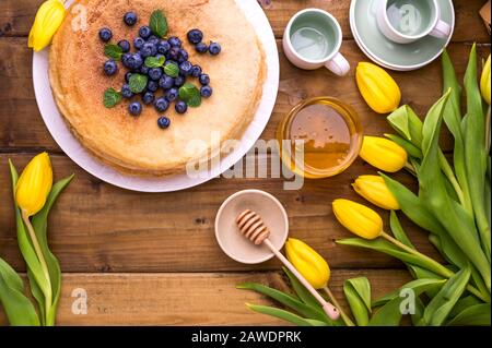 Große traditionelle russische Pfannkuchen mit Beeren und Honig auf einem Holztisch. Backen für den Frühlingsurlaub Shrovetide und einen Blumenstrauß gelber Tulpen. Kopierbereich. Stockfoto