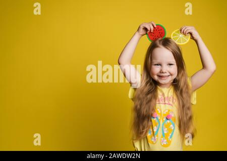 Fröhliches süßes kleines Mädchen, das ein Stück Obst über dem Kopf hält und Ohren mit einem unterhaltsamen mittleren Nahbereich macht Stockfoto