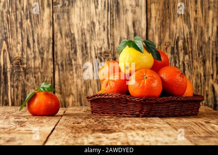 Frische Zitrusfrüchte, Mandarinen und Zitrone mit grünen Blättern im Korbkorb und auf einem alten Holztisch auf Holzgrund, Kopierraum. Stockfoto