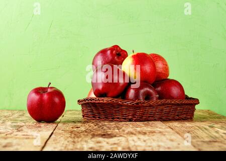 Rote reife Äpfel liegen in einem Korbkorb auf einem alten Holztisch auf hellgrünem Grund, Kopierraum. Stockfoto
