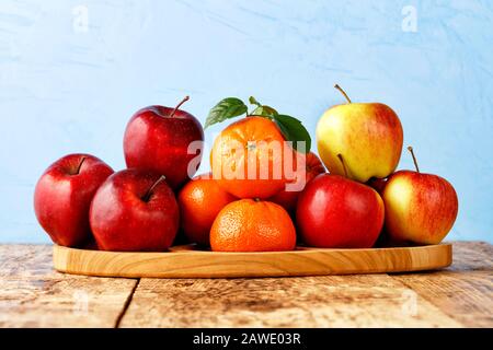 Rote reife Äpfel und Mandarinen mit grünen Blättern liegen auf einem Holztablett auf einem alten Holztisch auf hellblauem Grund, Kopierraum. Stockfoto