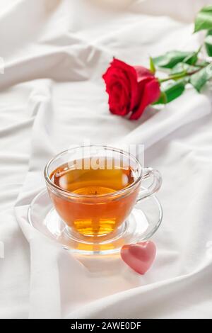 Wunderschöner romantischer Morgen mit einer Tasse Tee, herzförmiger Süßigkeiten und roter Rose auf weißem Bett Stockfoto