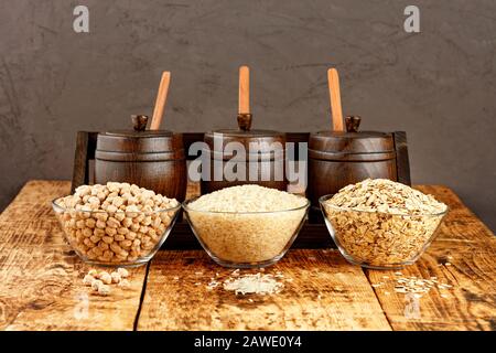 Kichererbsen, Reis und Haferbrei in Glasschüsseln vor dem Hintergrund kleiner Holzfässer zur Lagerung von Massenprodukten auf einem alten Holztisch. Stockfoto