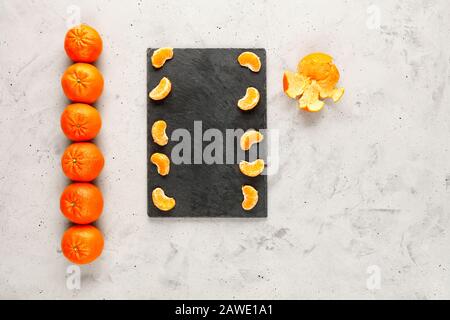 Helle und saftige Scheiben geschälten Mandarins liegen auf einem grauen Schiefer, der auf grauem Betongrund liegt, und es wird eine gleichmäßige Reihe ganzer orangefarbener Früchte angelegt Stockfoto