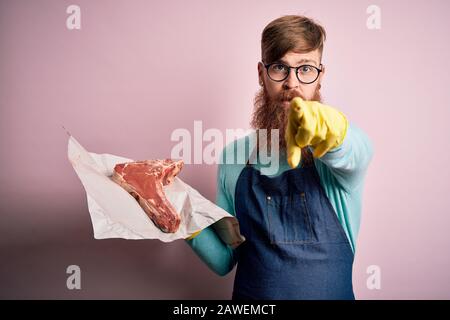 Rotkopf Irish Butcher man mit Bart, der rohes Rindersteak über rosafarbenem Hintergrund hält und mit dem Finger auf die Kamera und auf Sie zeigt, Handzeichen, positiv an Stockfoto