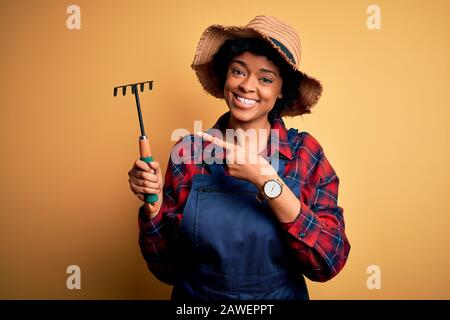 Junge afro-amerikanische afro-Bauernfrau mit geschweiften Haaren, die Schürze und Hut tragen und mit dem Rechen sehr glücklich zeigen, mit Hand und Finger Stockfoto