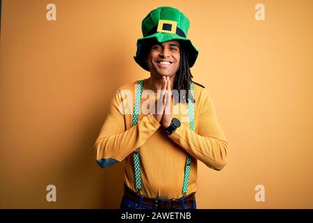 Junger afroamerikanischer Mann mit grünem Hut, der den Heiligen patricks Tag mit den Händen zusammen betet und um Vergebung bittet, lächelnd zuversichtlich. Stockfoto