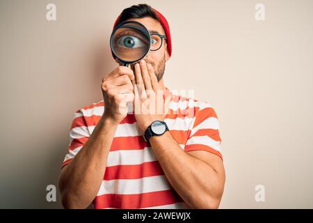 Junger Detektivmann, der mit der Hand schockiert aus versehen, Ausdruck der Angst, durch Lupe über den isolierten Hintergrundeöffnung blickt Stockfoto