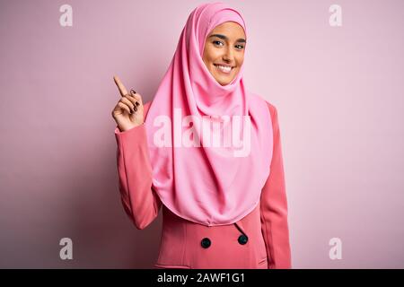 Junge, schöne Brunette-Geschäftsfrau mit pinkfarbenem muslimischem Hijab und Geschäftsjacke, die mit dem Finger Nummer eins zeigt und zeigt, während sie auf der Konferenz lächelt Stockfoto