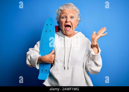 Senior schöne sportliche Frau, die Skate über isoliertem blauem Hintergrund hält, sehr glücklich und aufgeregt, Siegerausdruck feiert Siegesschreie Stockfoto