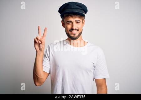 Junger Fahrer mit Bart und Hut, der über einem isolierten weißen Hintergrund steht, lächelt mit einem fröhlichen Gesicht, das an der Kamera winkelt und dabei ein Siegeszeichen macht. Taub Stockfoto