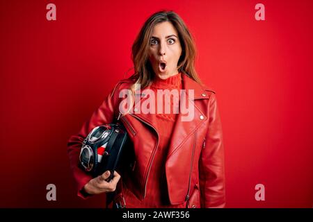 Schöne brünette Motorradfahrerin mit roter Jacke, die den Motorradhelm hält, erschrocken mit einem überraschenden Gesicht, Angst und aufgeregt mit Angst Stockfoto