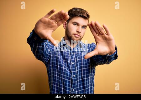 Junger blonder Geschäftsmann mit Bart und blauen Augen, der Hemd über gelbem Hintergrund trägt und mit Handflächen und Fingern Rahmen macht, Kameraperspektive Stockfoto