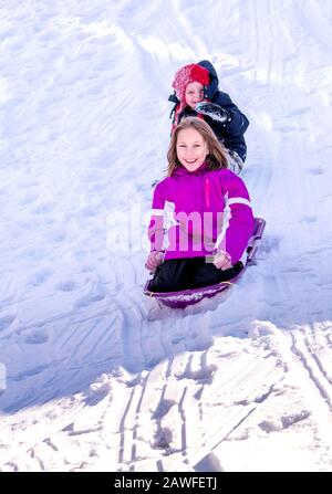 Zwei Kinder schlitten einen verschneiten Hügel hinunter, aber Schwester weiß nicht, dass ihr kleiner Bruder abgefallen ist Stockfoto