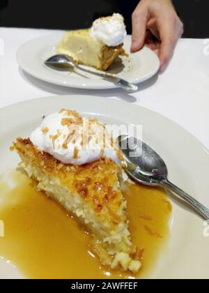 Ein Stück Kokosnusskuchen sitzt in einer Bourbon-Vanille-Glasur und wird mit gerösteter Kokosnuss und Schlagsahne bestockt. Eine Hand legt ein Stück Schlüsselkalk ab Stockfoto