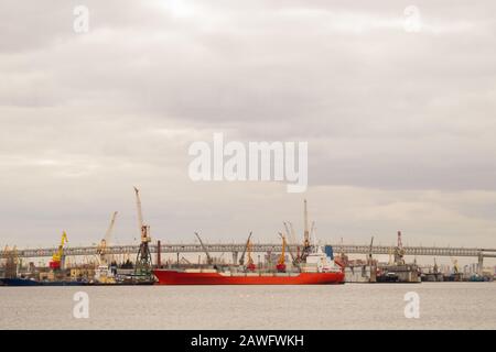 Rotes Frachtschiff im Hafen, Brücke über den Fluss mit Industriebetrieben, viele Industriekräne im Hafen Stockfoto