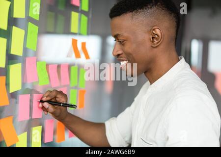 Lächelnder afroamerikanischer Geschäftsmann, der auf klebrigen Papieren schreibt Stockfoto