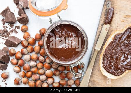 Hausgemachte vegane Haselnussschokolade auf Sauerteigbrot und in einem kilner Krug, mit ungeschelten Haselnüssen und Brocken dunkler Schokolade Stockfoto