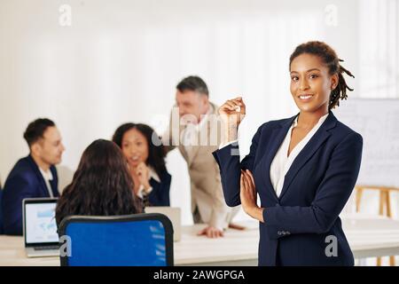 Hübsche junge Geschäftsfrau mit wunderschönem Lächeln und ihren Kollegen, die im Hintergrund über das Projekt diskutieren Stockfoto