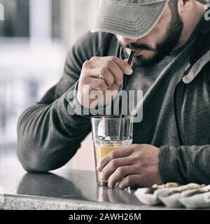Starker junger aktiver Mann mit Bartkappe und Sporthoodie, der Orangensaft mit Strohhalm für Getränke trinkt Stockfoto