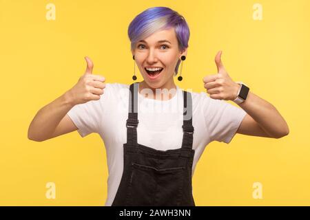 Wie! Porträt einer optimistischen fröhlichen Hüftfrau mit violett gefärbten kurzen Haaren in Denim-Overalls, die doppelte Daumen nach oben zeigen und Wow Ja sagen, weit offen Stockfoto