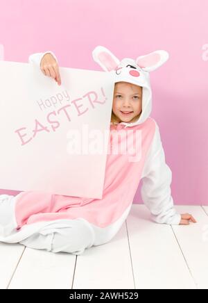 Hübsches blondes Mädchen mit gemütlichem Kaninchen-Kostüm und weißem Schild mit den Worten Happy easter darauf posiert im Studio und ist glücklich Stockfoto