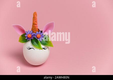 Niedliche Frühlings-Osterkarte mit einem Einhorn-Ei mit Federblumen und Horn mit Kopffarbe auf pastellpinkem Hintergrund Stockfoto